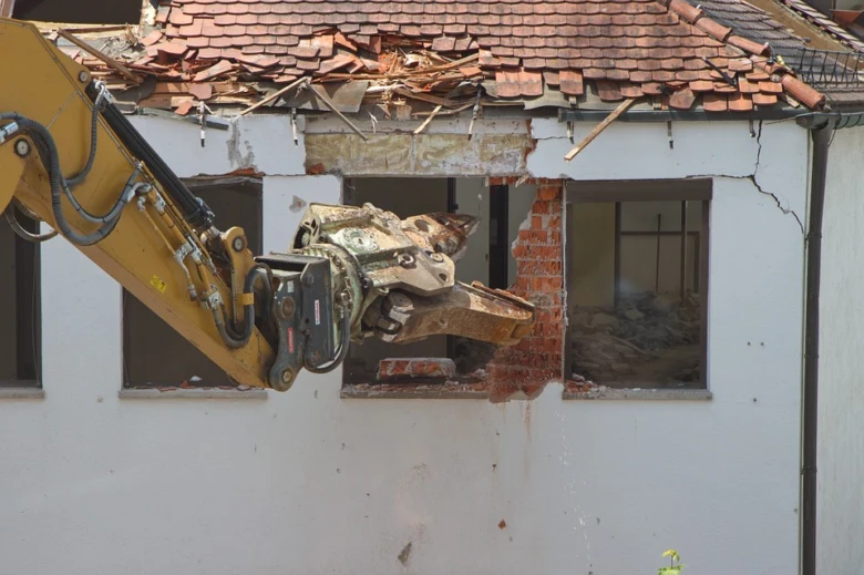 Žlutý bagr rozbíjí zeď při demolicích domu.