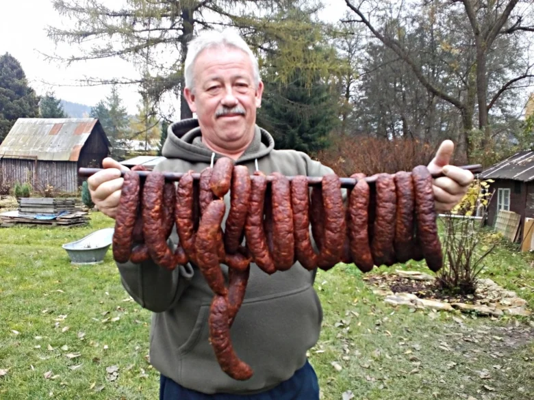 Muž drží v rukou řadu uzených klobás na tyči venku.