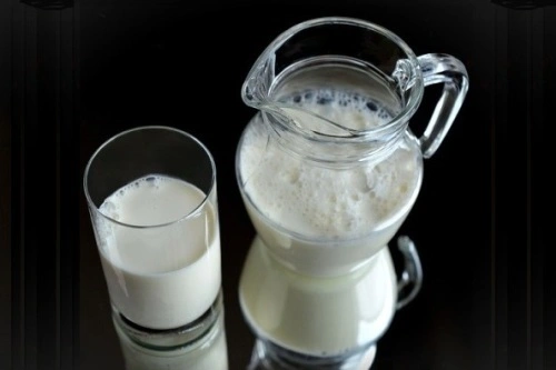 Sklenička mléka vedle džbánu s mlékem na tmavém pozadí.