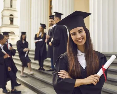 Usměvavá absolventka s diplomem na schodech univerzity.