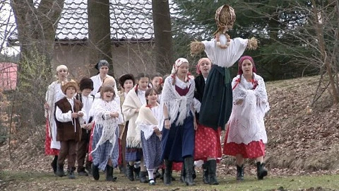 Skupina lidí v lidových kostýmech na venkově s kukuřičnou panenou.