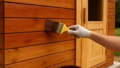 Ruka v rukavici nanáší lak na dřevěné stěny sauny štětcem.