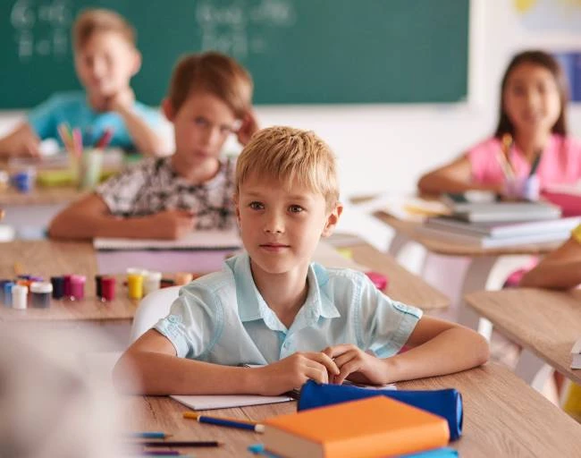 Školák sedí u stolu v třídě s ostatními dětmi.