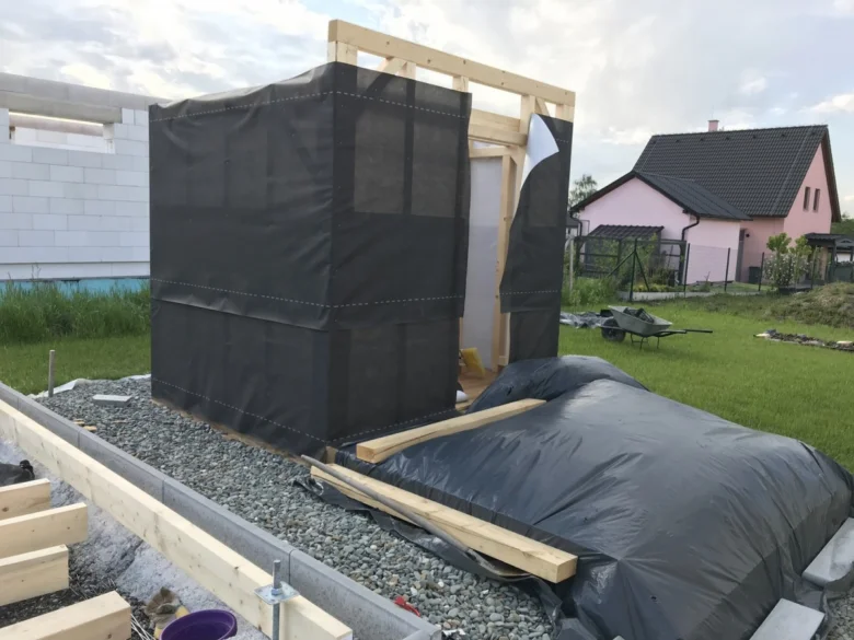 Sauna v stavbě s dřevěným rámem a černou fólií na pozadí trávníku.